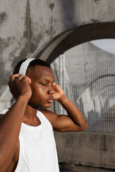 Calm African American male with eyes closed in wireless headphones listening to songs while training outdoors - ADSF46474