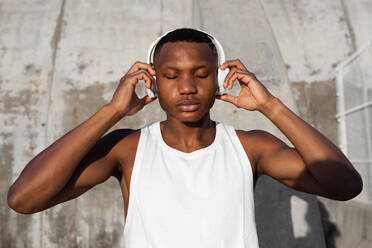 Calm African American male with eyes closed in wireless headphones listening to songs while training outdoors - ADSF46476