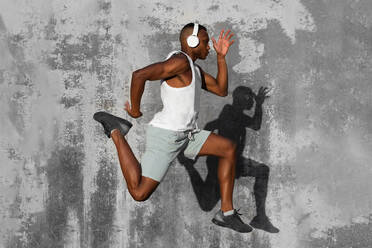 Full body side view of concentrated African American runner listening to music in headphones while jumping above ground during jogging training - ADSF46477