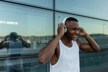 Cheerful African American male athlete smiling brightly while listening to music in wireless headphones during fitness training - ADSF46478