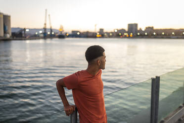 Young ethnic male traveler standing behind rippled sea under sky in twilight and looking away - ADSF46480