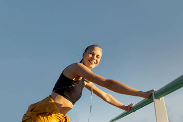 Young cheerful athletic caucasian woman leaning on banister outdoors and looking at camera after working out - ADSF46482