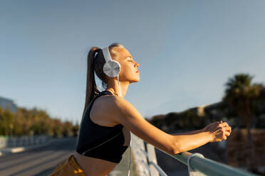 Young athletic caucasian leaning on banister outdoors, listening to music and relaxing with closed eyes after working out - ADSF46484