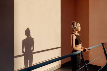 Young woman leaning on banister and listening to music using phone after working out outdoors at sunset - ADSF46493