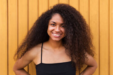 Delighted African American female with Afro hairstyle standing near wooden wall and sincerely smiling at camera - ADSF46504