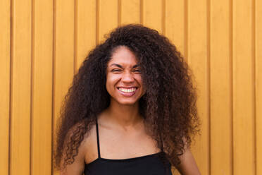 Delighted African American female with Afro hairstyle standing near wooden wall and sincerely smiling at camera - ADSF46505