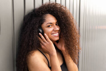 Lovely African American female with long curly hair and headphones in city and looking at camera - ADSF46509