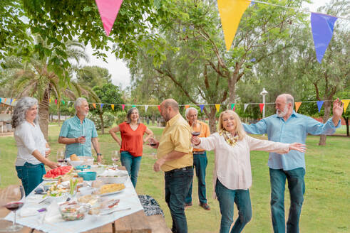 Group of middle aged male and female friends in casual clothes dancing near table with food while celebrating birthday in park - ADSF46515