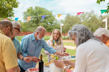 Group of middle aged men and women eating food and drinking wine during outdoor party in yard - ADSF46517