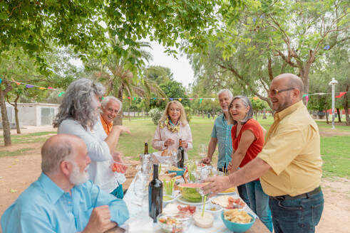 Group of middle aged men and women in casual clothes eating food and drinking wine during outdoor party in park - ADSF46520