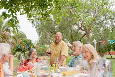 Positive bald man in orange shirt standing near table with glass of red wine and making toast while celebrating birthday in park - ADSF46529