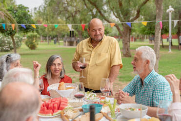 Smiling bald man in orange shirt standing near table with glass of red wine and making toast while celebrating birthday in park - ADSF46531