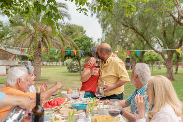 Middle aged couple hugging and kissing while celebrating occasion with friends in green park - ADSF46532