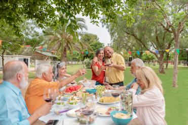 Middle aged man and woman hugging and clinking glasses while celebrating anniversary with friends in green park on sunny day - ADSF46535