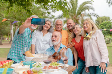 Group of cheerful middle aged men and women standing near table with assorted food and wine while taking self portrait on smartphone - ADSF46541