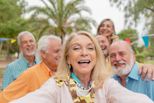 Smiling middle aged woman with blond hair taking self portrait with friends during outdoor celebration in park - ADSF46543