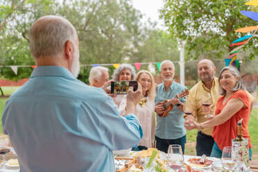Group of middle aged people listening to bearded man playing on ukulele and singing during outdoor celebration and filming video on smartphone - ADSF46544