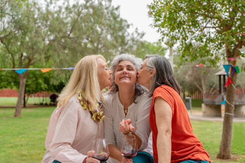 Happy middle aged women in casual clothes sitting on grass with glasses of red wine and kissing girlfriend to cheeks in park decorated for celebration - ADSF46545