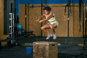 Full body of determined African American sportswoman in activewear doing box jumps with arms outstretched forward during fitness workout in gym - ADSF46552