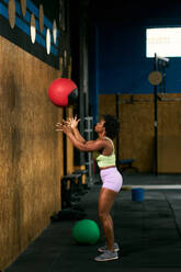 Full body side view of sportive African American female athlete crouching and throwing red medicine ball against wall during training in gym - ADSF46553