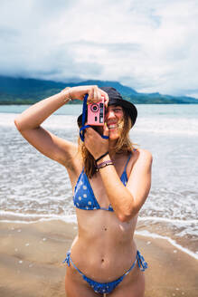 Crop young female tourist in bikini and hat standing on wet sandy Uvita beach while taking photo on film vintage camera during trip through Costa Rica - ADSF46560