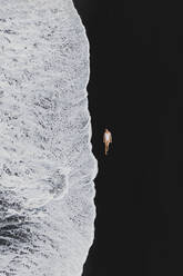 Aerial view of a girl lay down on a black sand beach in Lanzarote, Canary Islands, Spain. - AAEF19798