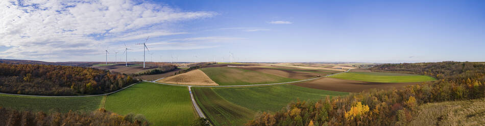 Aerial drone panoramic view of agricultural fields bordered by woods with autumn colours and wind turbines in the background under a semi-clear sky in Lower Austria. - AAEF20044