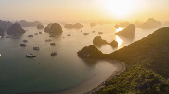 Panoramic aerial view of sunset in the hazy horizon of Halong Bay, showing a beach, multiple boats and small islands scattered across the bay, Vietnam. - AAEF20105