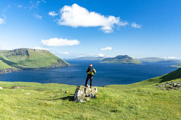 Hiker standing on top of rock contemplating the fjord in summer, Nordradalur, Streymoy Island, Faroe Islands, Denmark, Europe - RHPLF26090