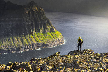 One man admiring the majestic cliffs along a fjord standing on top of mountain during a hike, Vidoy Island, Faroe Islands, Denmark, Europe - RHPLF26095