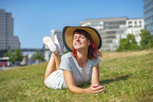 Smiling woman wearing sun hat relaxing on grass with eyes closed - BFRF02432