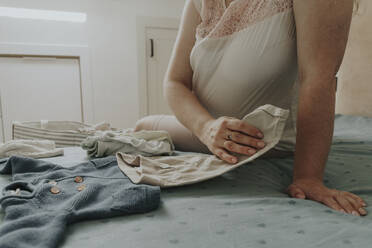 Pregnant woman choosing baby clothing on bed at home - OSF01950