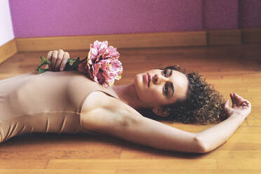 Contemplative woman holding flower and lying on hardwood floor - JSMF02863