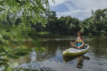Woman meditating on paddleboard in lake - YTF01028
