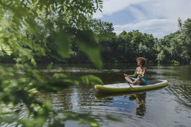 Woman sitting and meditating on paddleboard in lake - YTF01029