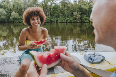 Lächelnde Frau isst Wassermelone mit Freund auf Paddleboard in See - YTF01035