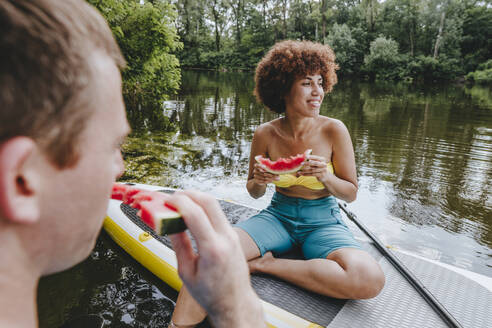 Nachdenkliche Frau isst Wassermelone mit einem Freund auf einem Paddelbrett im See - YTF01037