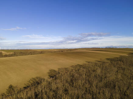 Aerial drone view in winter of agricultural fields and countryside with scattered clouds against a blue sky with wind turbines in the distance, Spannberg, Lower Austria, Austria. - AAEF21393