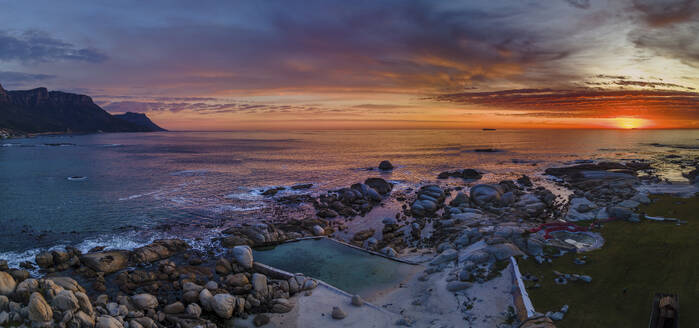 Panoramic aerial view of Maiden’s Cove Tidal Pool at dramatic sunset with Camps Bay in background, Cape Town, South Africa. - AAEF21419