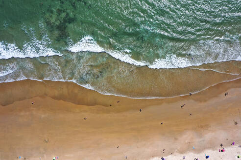 Aerial view of the blue ocean with the white waves and the brown sand with people on it in the town of Conil de la Frontera on Playa La Fontanilla in Cádiz, Andalusia, Spain. - AAEF21732