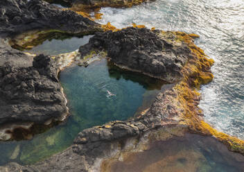 Aerial view of a person swimming in a natural rock pool along the coastline with cliffs at Ponta dos Mosteiros, Ilha de Sao Miguel, Azores, Portugal. - AAEF21791