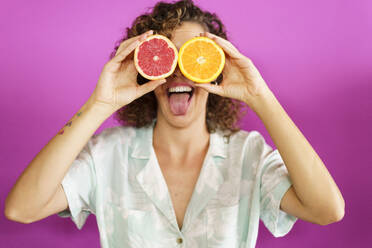 Playful woman covering eyes with slice of grapefruit and orange against magenta background - JSMF02883