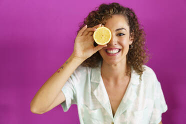 Glückliche Frau, die eine Zitronenscheibe über das Auge hält, vor magentafarbenem Hintergrund - JSMF02886