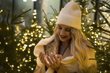 Smiling woman holding string lights near Christmas trees - YBF00097