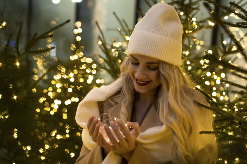 Lächelnde Frau mit Lichterkette in der Nähe von Weihnachtsbäumen - YBF00097