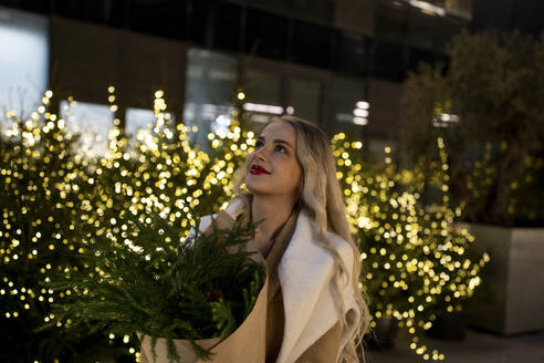 Lächelnde Frau mit Blumenstrauß in der Nähe der Weihnachtsbeleuchtung - YBF00098