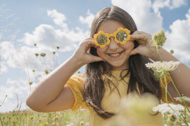 Smiling girl with flower sunglasses pulling cheeks in field - OSF01970