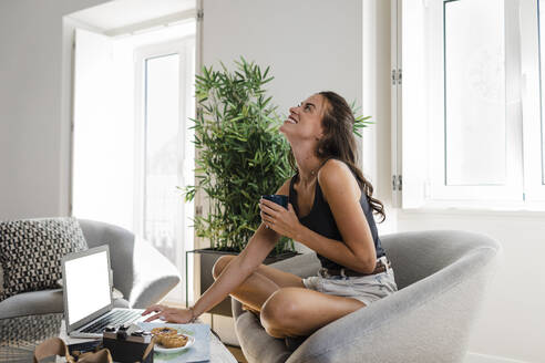 Glückliche junge Frau mit Kaffeetasse und Laptop auf einem Sessel sitzend - DCRF01664