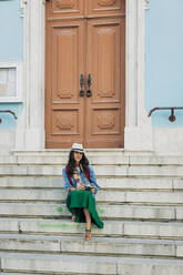 Lächelnde Frau mit Hut sitzt auf den Stufen vor der Tür - DCRF01723