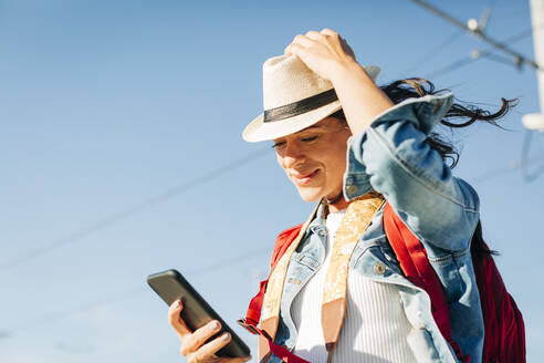 Lächelnde junge Frau mit Hut und Smartphone unter dem Himmel - DCRF01729